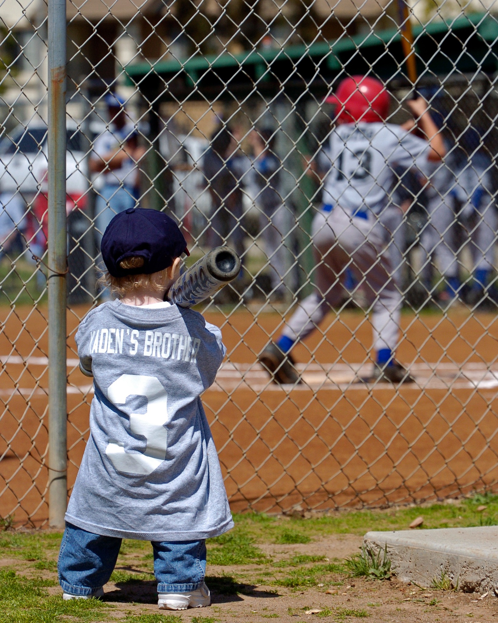 Young child watching a baseball game through the fence with a bat on his shoulder